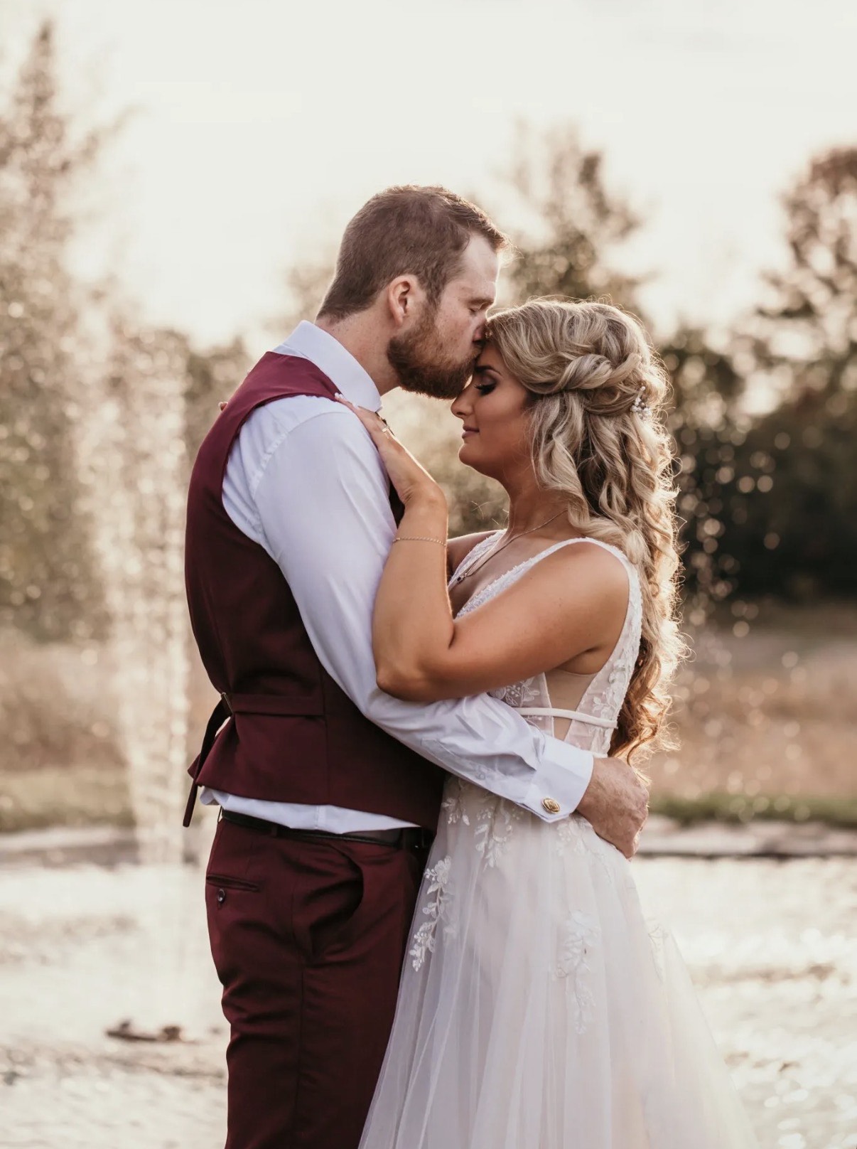 Groom kissing the bride's forehead by the estate pond fountain